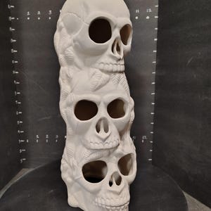 May include: A white ceramic sculpture of three stacked skulls. The skulls have large eye sockets and detailed teeth. The sculpture is decorated with leaf-like patterns between the skulls. The piece is displayed on a black surface.