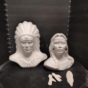 May include: Two white ceramic busts depicting individuals with traditional headwear, including a figure with a large feathered headdress and a figure with braided hair and a headband, accompanied by two small separate white ceramic feather pieces.