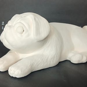 May include: White ceramic pug figurine, lying down with its head turned to the side. The figurine is detailed with realistic features, including wrinkles and fur.