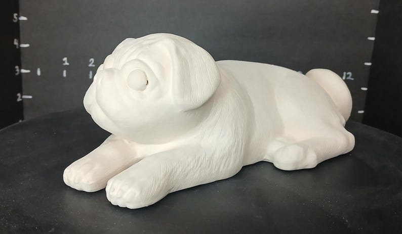 May include: White ceramic pug figurine, lying down with its head turned to the side. The figurine is detailed with realistic features, including wrinkles and fur.