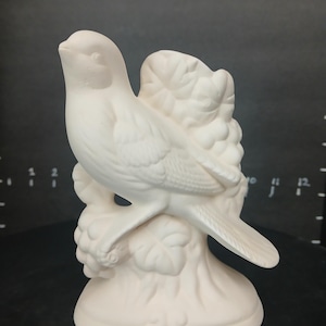 May include: White ceramic bird figurine perched on a branch with grapes. The bird is facing to the right and has its wings spread. The figurine is ready to be painted.