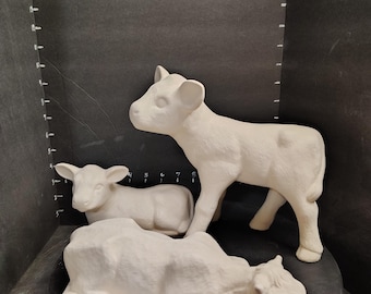 May include: Three unpainted ceramic animal figurines consisting of a standing calf, a small lamb lying down, and a larger cow lying down. The standing calf measures approximately 8 inches in height against a black backdrop with a numbered scale.