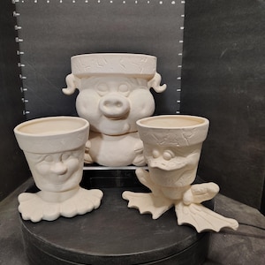 May include: Three unpainted ceramic planters. One is a pig with a large snout and ears. The other two planters have faces with a collar and webbed feet. The planters are on a black surface.