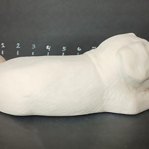 May include: White ceramic pug figurine in a sleeping position. The figurine is detailed with realistic features, including wrinkles and folds on the face and body.
