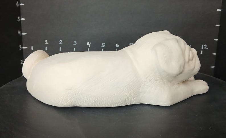 May include: White ceramic pug figurine in a sleeping position. The figurine is detailed with realistic features, including wrinkles and folds on the face and body.