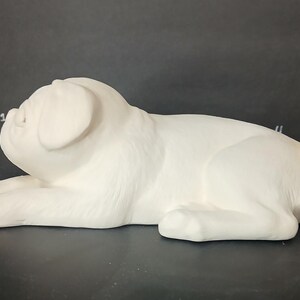 May include: White ceramic pug figurine lying down with its head turned to the side. The figurine is unpainted and ready to be decorated.