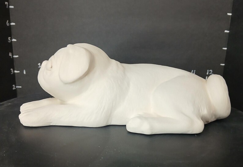 May include: White ceramic pug figurine lying down with its head turned to the side. The figurine is unpainted and ready to be decorated.
