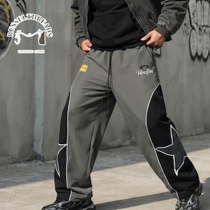 May include: Grey and black track pants featuring a drawstring waist, a small yellow rectangular patch on the right hip, and a white embroidered logo on the left thigh. The legs have large black side panels with white star outlines.