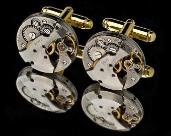 Puede incluir: Un par de gemelos únicos que presentan los intrincados mecanismos internos de un reloj. Los componentes metálicos plateados, incluyendo engranajes y joyas, están colocados sobre un respaldo dorado. Los gemelos se reflejan en una superficie negra.