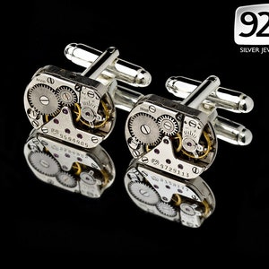 Puede incluir: Un par de gemelos de plata con un diseño de mecanismo de relojería detallado y expuesto. Los gemelos están hechos de plata y tienen un acabado pulido. El mecanismo de relojería es visible a través de la cara transparente de los gemelos. Los gemelos son un accesorio único y elegante para cualquier hombre.