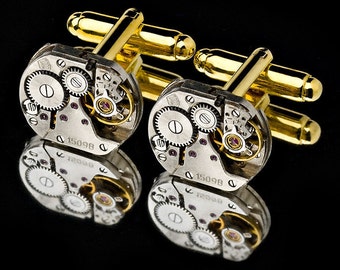 May include: A pair of gold cufflinks with a silver watch movement design. The cufflinks feature intricate gears and a small ruby-colored jewel. The cufflinks are reflected in a black surface.