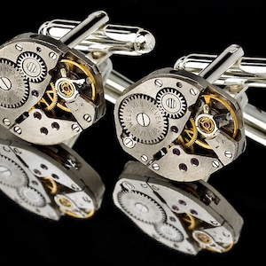 Puede incluir: Un par de gemelos de plata con un diseño detallado de mecanismo de relojería expuesto. Los gemelos presentan un esquema de color plateado y dorado con engranajes visibles y una inscripción 'Seven Jewels'.