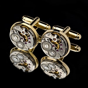 May include: A pair of gold-toned cufflinks with a watch mechanism design. The round cufflinks feature visible gears, jewels, and intricate details, reflecting light against a black background. The cufflinks have a polished finish.