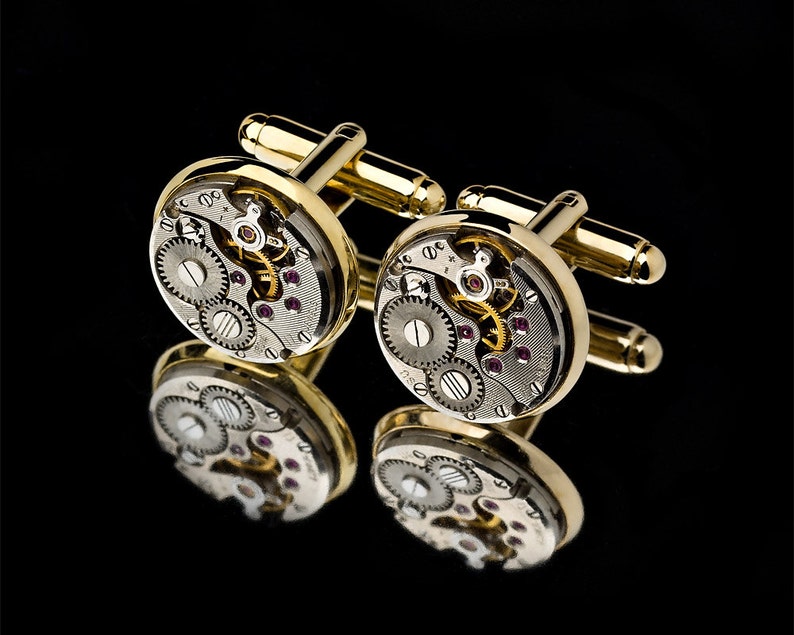 May include: A pair of gold-toned cufflinks with a watch mechanism design. The round cufflinks feature visible gears, jewels, and intricate details, reflecting light against a black background. The cufflinks have a polished finish.