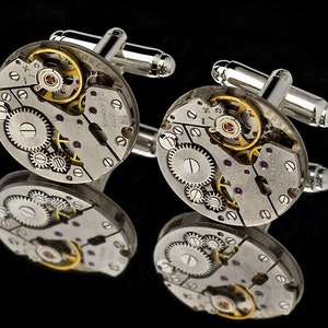 May include: A pair of silver cufflinks with a circular design featuring exposed watch gears and a gold-colored winding mechanism.