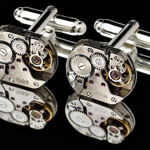 Puede incluir: Un par de gemelos de plata con un diseño único que presenta engranajes de reloj expuestos y un acabado plateado. Los gemelos tienen una forma circular y están decorados con detalles intrincados.
