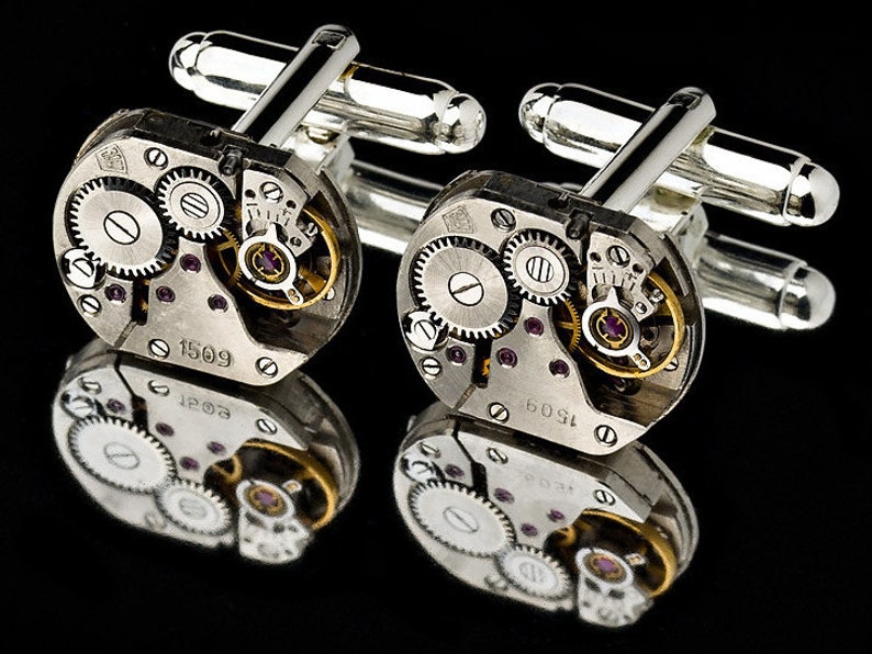 May include: A pair of silver cufflinks with a unique design featuring exposed watch gears and a silver finish. The cufflinks have a circular shape and are decorated with intricate details.