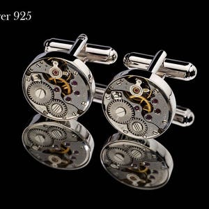 May include: Silver 925 cufflinks with a watch mechanism design. The silver cufflinks feature intricate gears, ruby-colored jewels, and a polished finish. The cufflinks are displayed on a reflective black surface.