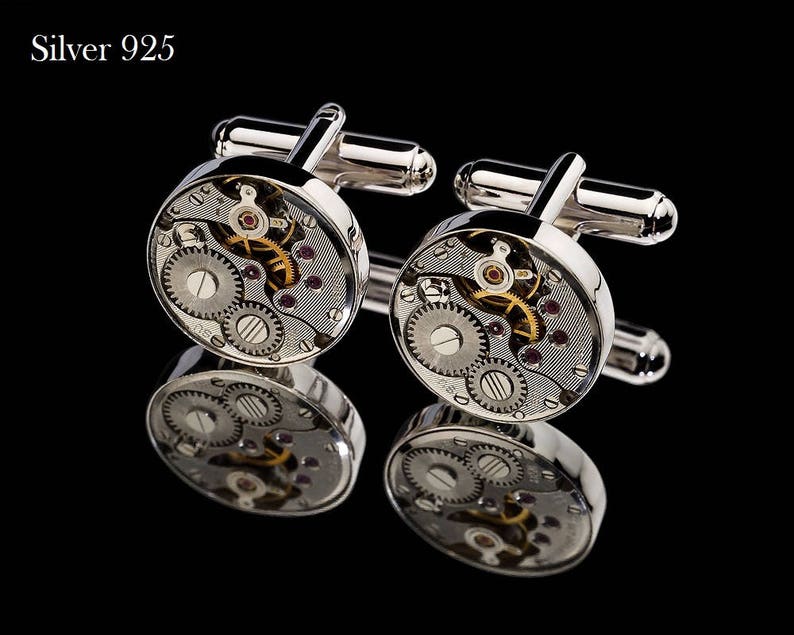 May include: Silver 925 cufflinks with a watch mechanism design. The silver cufflinks feature intricate gears, ruby-colored jewels, and a polished finish. The cufflinks are displayed on a reflective black surface.