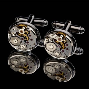May include: A pair of silver cufflinks with a watch mechanism design. The cufflinks feature visible gears, jewels, and intricate details, reflecting light against a black background. The design includes silver bars for fastening.