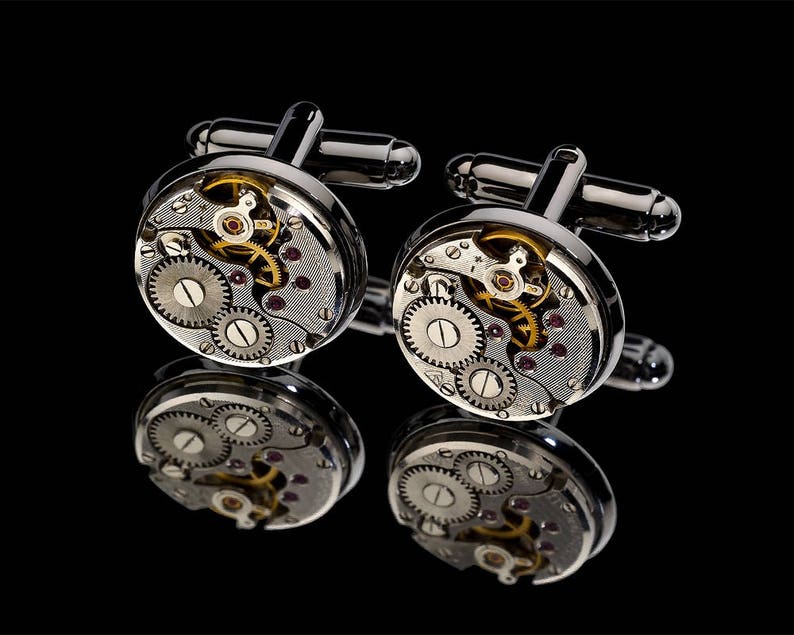 May include: A pair of silver cufflinks with a watch mechanism design. The cufflinks feature visible gears, jewels, and intricate details, reflecting light against a black background. The design includes silver bars for fastening.