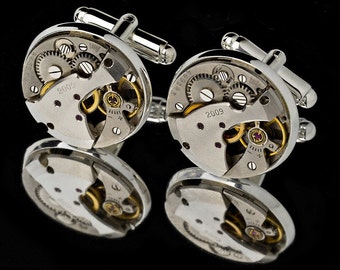 Puede incluir: Un par de gemelos de plata con mecanismos de reloj expuestos. Los gemelos presentan un acabado plateado y engranajes dorados. Los gemelos están grabados con el año 2009.