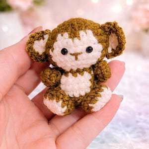 May include: A handmade crochet monkey doll in shades of brown and white. The small stuffed animal has a detailed design with visible stitches, black eyes, and a cute expression. The monkey is held in a person's hand.