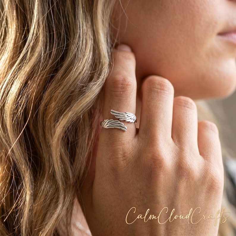 Puede incluir: Un anillo plateado con dise&ntilde;o de alas. El anillo envuelve el dedo, con cada ala extendi&eacute;ndose desde lados opuestos. El anillo se lleva en una mano, con cabello rubio largo y ondulado en el fondo. El texto "Calm Cloud Crafts" es visible.