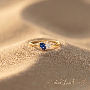 May include: A gold ring with a split design, featuring a pear-shaped blue gemstone on one side and a clear gemstone on the other. The ring is set against a sandy background, with the text "Calm Cloud Crafts" in the lower right corner.