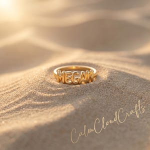 May include: A gold ring spelling out the name "MEGAN" in capital letters. The ring is set on sand, with the words "Calm Cloud Crafts" in cursive script in the lower right corner.
