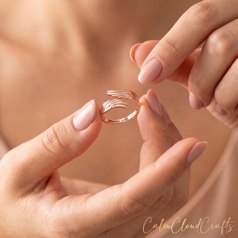 Puede incluir: Anillo de oro rosa con un dise&ntilde;o abierto con alas de &aacute;ngel estilizadas. El anillo est&aacute; grabado con las palabras "always with you". El anillo se sostiene entre dos manos.