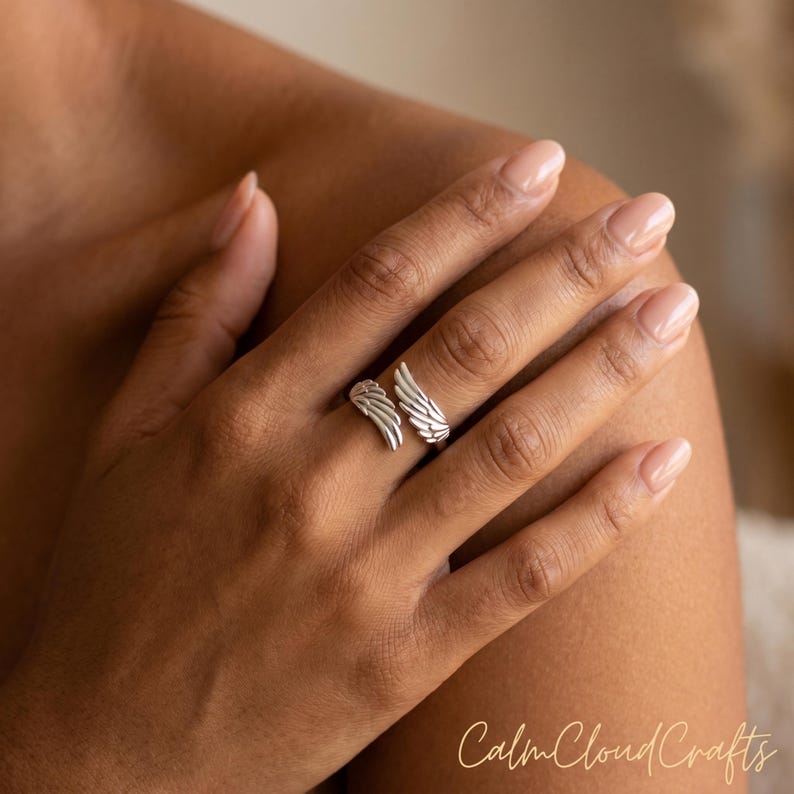 Puede incluir: Un anillo plateado con dise&ntilde;o de alas. El anillo envuelve el dedo, con un ala a cada lado. El anillo est&aacute; en una mano con u&ntilde;as cuidadas. Las palabras "Calm Cloud Crafts" est&aacute;n en la esquina inferior derecha.