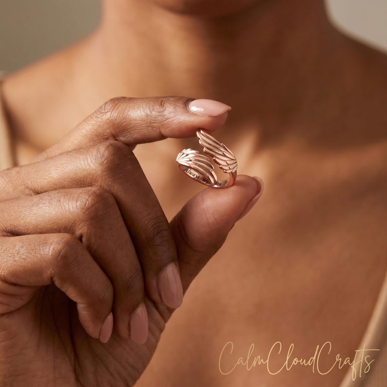Puede incluir: Anillo de color oro rosa con un dise&ntilde;o abierto con detalles en forma de alas. El anillo tiene la palabra "Always" grabada. El anillo est&aacute; sujeto entre los dedos. El dise&ntilde;o del anillo recuerda a las alas de un &aacute;ngel.