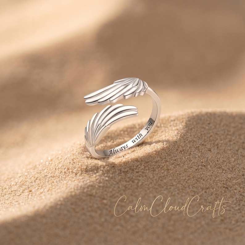 Puede incluir: Un anillo plateado con dise&ntilde;o de ala. El anillo es abierto, con las puntas de las alas superpuestas. Las palabras "Always with you" est&aacute;n grabadas en el anillo. El anillo est&aacute; sobre una superficie arenosa.