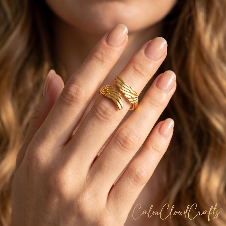 Puede incluir: Un anillo dorado con dise&ntilde;o de ala. El anillo se lleva en un dedo, con las alas envolviendo el dedo. El anillo es de metal brillante y tiene una textura detallada. El anillo es una joya.