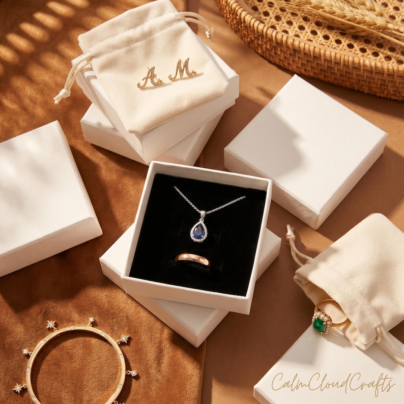 Puede incluir: Una variedad de joyas, que incluyen un collar con colgante de zafiro, un anillo de oro y una pulsera con detalles en forma de estrella. Las joyas se exhiben en cajas blancas y peque&ntilde;as bolsas con cord&oacute;n. El texto "Calm Cloud Crafts" es visible.