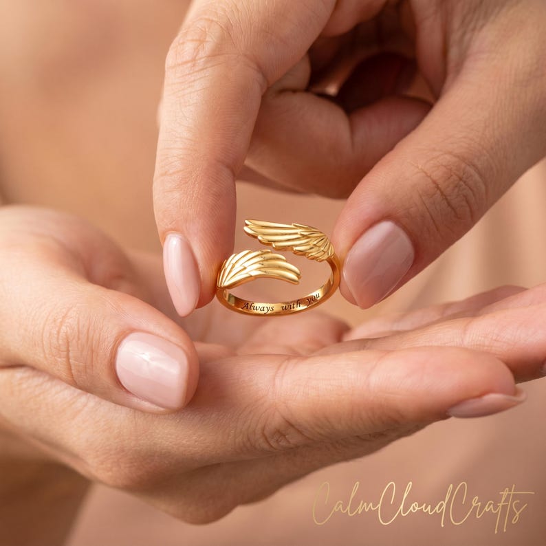 Puede incluir: Un anillo dorado con un dise&ntilde;o abierto, con detalles en forma de alas en cada extremo. El anillo tiene la inscripci&oacute;n "Always with you". El anillo est&aacute; sostenido entre dos manos.