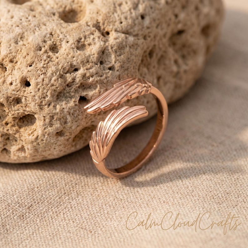 Puede incluir: Un anillo de oro rosa con dise&ntilde;o de pluma. El anillo presenta un dise&ntilde;o abierto con dos extremos en forma de pluma que se curvan alrededor del dedo. El anillo est&aacute; sobre un fondo neutro con el texto "Calm Cloud Crafts".