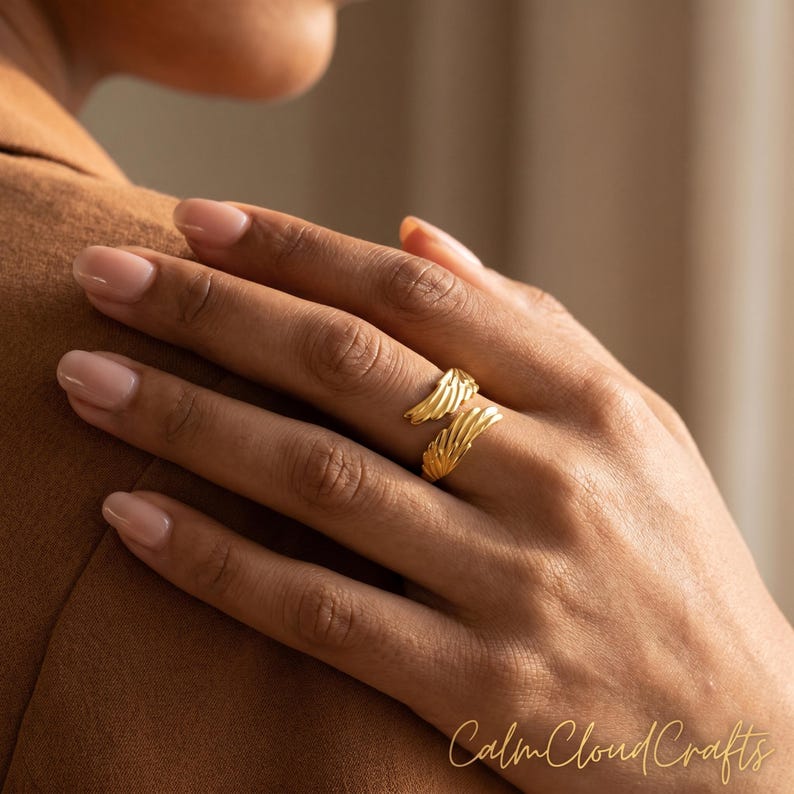 Puede incluir: Un anillo dorado con un dise&ntilde;o en capas en forma de ala se lleva en el dedo de una persona. El anillo es el punto focal, con la mano y el brazo de la persona en el encuadre. El texto "Calm Cloud Crafts" es visible.