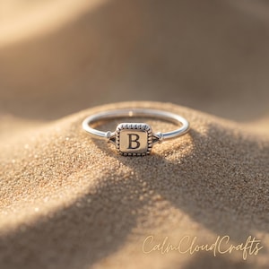 May include: A silver ring with a square, textured centerpiece featuring the letter "B". The ring is set against a sandy background, with the ring's band appearing thin and delicate. The ring is a piece of jewellery.