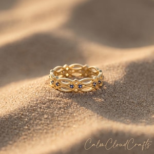 Puede incluir: Un anillo dorado con un diseño calado con formas ovaladas repetidas y pequeñas piedras preciosas de color azul oscuro. El anillo está sobre arena. El texto "Calm Cloud Crafts" está escrito en cursiva en la parte inferior.
