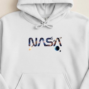 Könnte beinhalten: Weißer Kapuzenpullover mit Fronttasche und Kordelzug. Das Wort "NASA" ist in einem Weltraum-Design mit einer Galaxie und Planeten auf die Brust gedruckt.