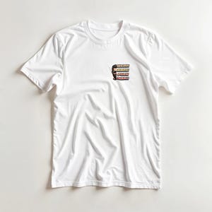 Op de afbeelding: Wit T-shirt met een kleine afbeelding op de linkerbovenborst. De afbeelding toont een gestileerd gezicht en de woorden "WORK HARD PLAY HARD" verticaal gestapeld in een retro lettertype. Het shirt heeft korte mouwen en een ronde hals.