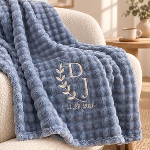 May include: A dusty blue textured waffle-knit throw blanket featuring an embroidered design with the letters D and J, a laurel leaf branch, and the date 11.29.2026 stitched in a light cream thread color across the front surface of the fabric.