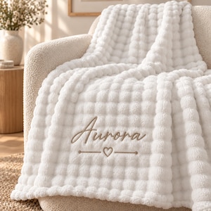 May include: A white plush throw blanket with a textured bubble pattern. The blanket features the name Aurora embroidered in a light brown script font, positioned above a small heart icon flanked by two horizontal lines in the same color.
