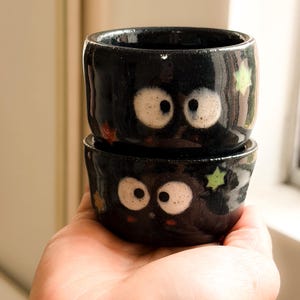 Puede incluir: Dos cuencos de cerámica negros apilados, cada uno con un diseño de cara de dibujos animados. Las caras tienen ojos blancos grandes con pupilas negras y pequeños detalles rojos. Se ven detalles de estrellas verdes y naranjas.