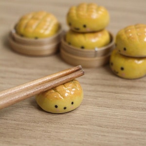 May include: A set of miniature yellow bread-shaped figures with small black eyes and pink cheeks, featuring a cross-hatched top pattern. One figure serves as a rest for a pair of wooden chopsticks, while others are placed in small bamboo containers.