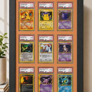 May include: Framed display of twelve Pok&eacute;mon trading cards, featuring Charizard, Pikachu, Gengar, Rayquaza, Lugia, Mewtwo, Umbreon, Espeon, and Gyarados. The cards are encased in protective holders and arranged in a black frame with an orange background.