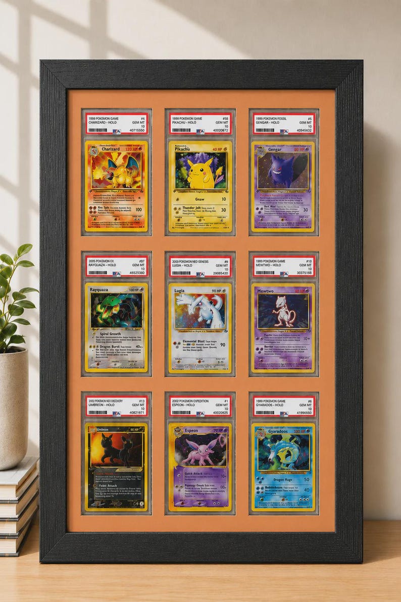 May include: Framed display of twelve Pok&eacute;mon trading cards, featuring Charizard, Pikachu, Gengar, Rayquaza, Lugia, Mewtwo, Umbreon, Espeon, and Gyarados. The cards are encased in protective holders and arranged in a black frame with an orange background.