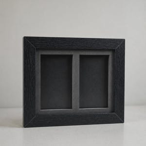 May include: A black, double-aperture picture frame. The frame has a textured wood grain appearance and a rectangular shape. The two openings are rectangular and have a dark gray background. The frame is designed to hold two photos.